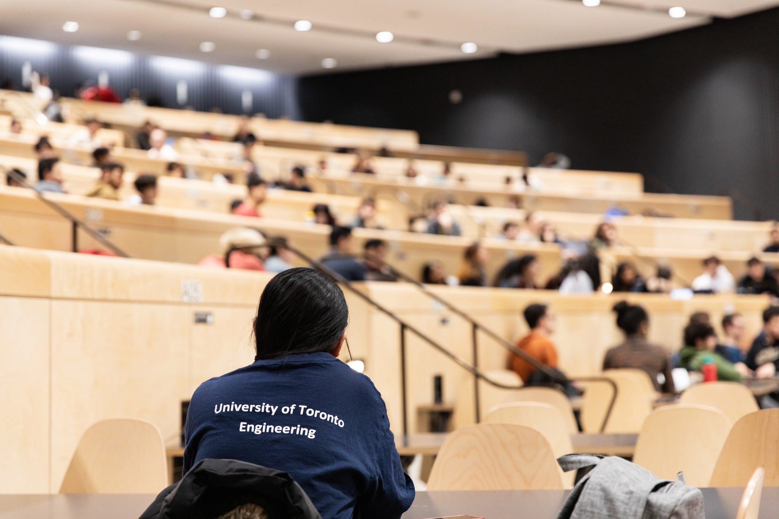 Photo shows a lecture hall full of students. It highlights a student wearing a U of T Engineering t-shirt.